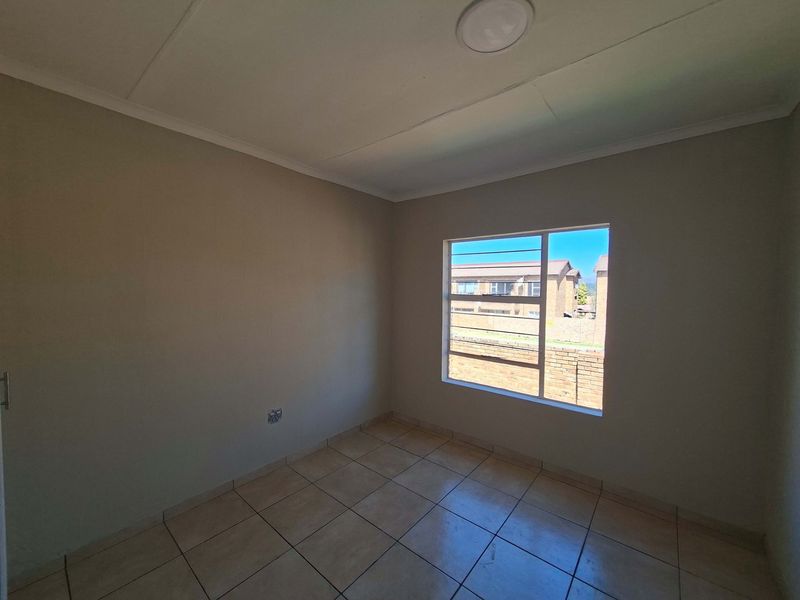 A clean, empty bedroom featuring neutral wall tones, tiled flooring, and a large window providing natural light and a view of the surrounding complex.