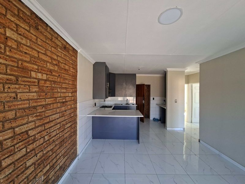 Open plan kitchen and living area featuring a striking exposed brick wall, modern tiled flooring, and contemporary dark cabinetry with a breakfast bar.