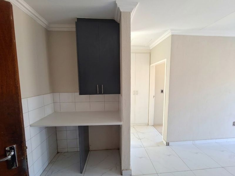 A clean, empty kitchen area featuring modern white tiled flooring, built-in dark cabinetry, and a tiled splashback, leading into a hallway with built-in storage.