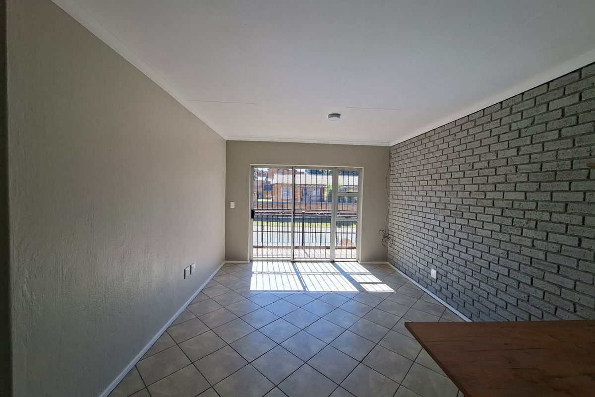 An empty living room featuring tiled flooring, an exposed brick feature wall, and a sliding glass door with a security gate leading outside.