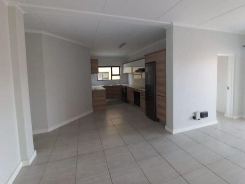 An open-plan living and kitchen area featuring neutral tiled flooring, modern wood-finish cabinetry, and integrated kitchen appliances in a clean, vacant space.