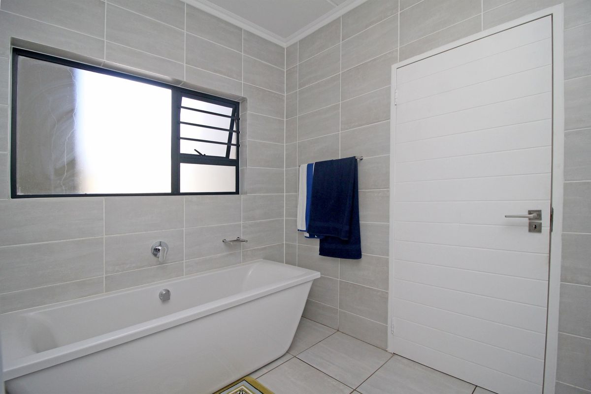 A clean, modern bathroom featuring neutral wall and floor tiles, a white bathtub, and a frosted glass window for privacy.