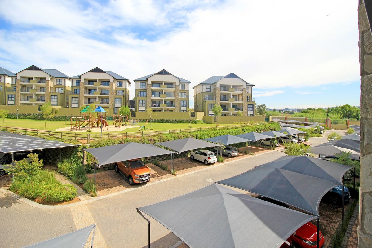 Elevated view of the apartment complex grounds featuring paved parking with shade cloth carports, landscaped gardens, and a communal children's playground area.