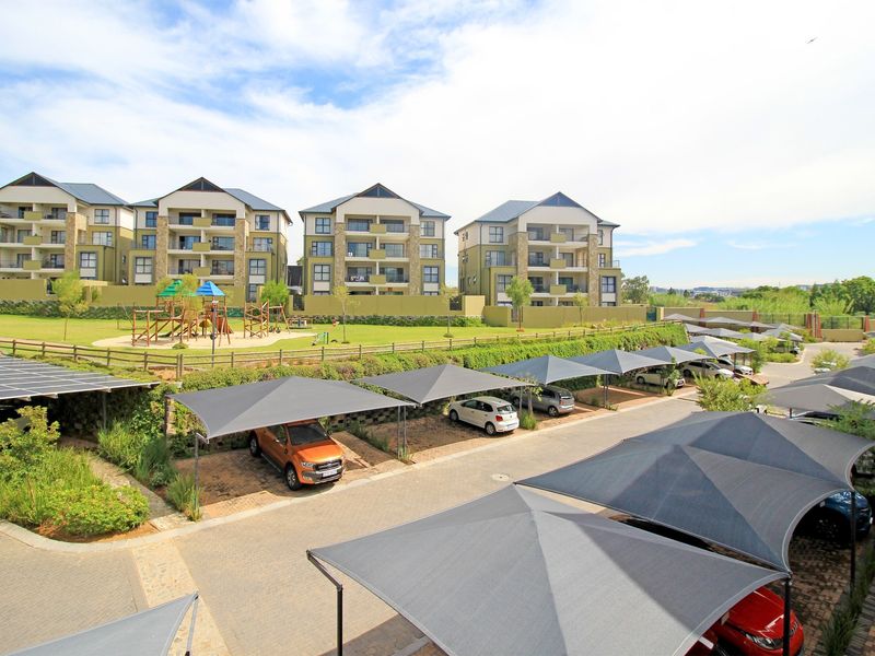 Elevated view of the apartment complex grounds featuring paved parking with shade cloth carports, landscaped gardens, and a communal children's playground area.