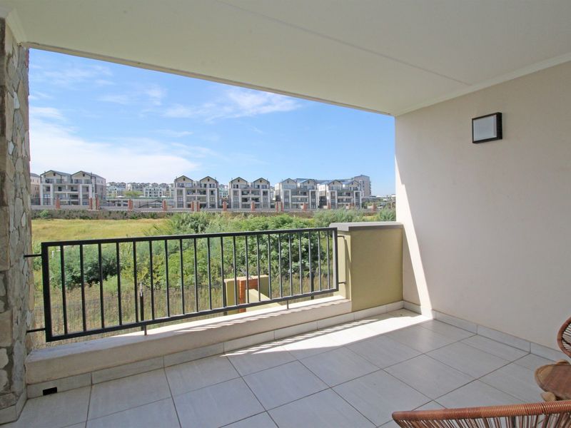 A spacious covered balcony featuring tiled flooring, a metal balustrade, and a decorative stone-clad pillar, offering open views of the surrounding estate.