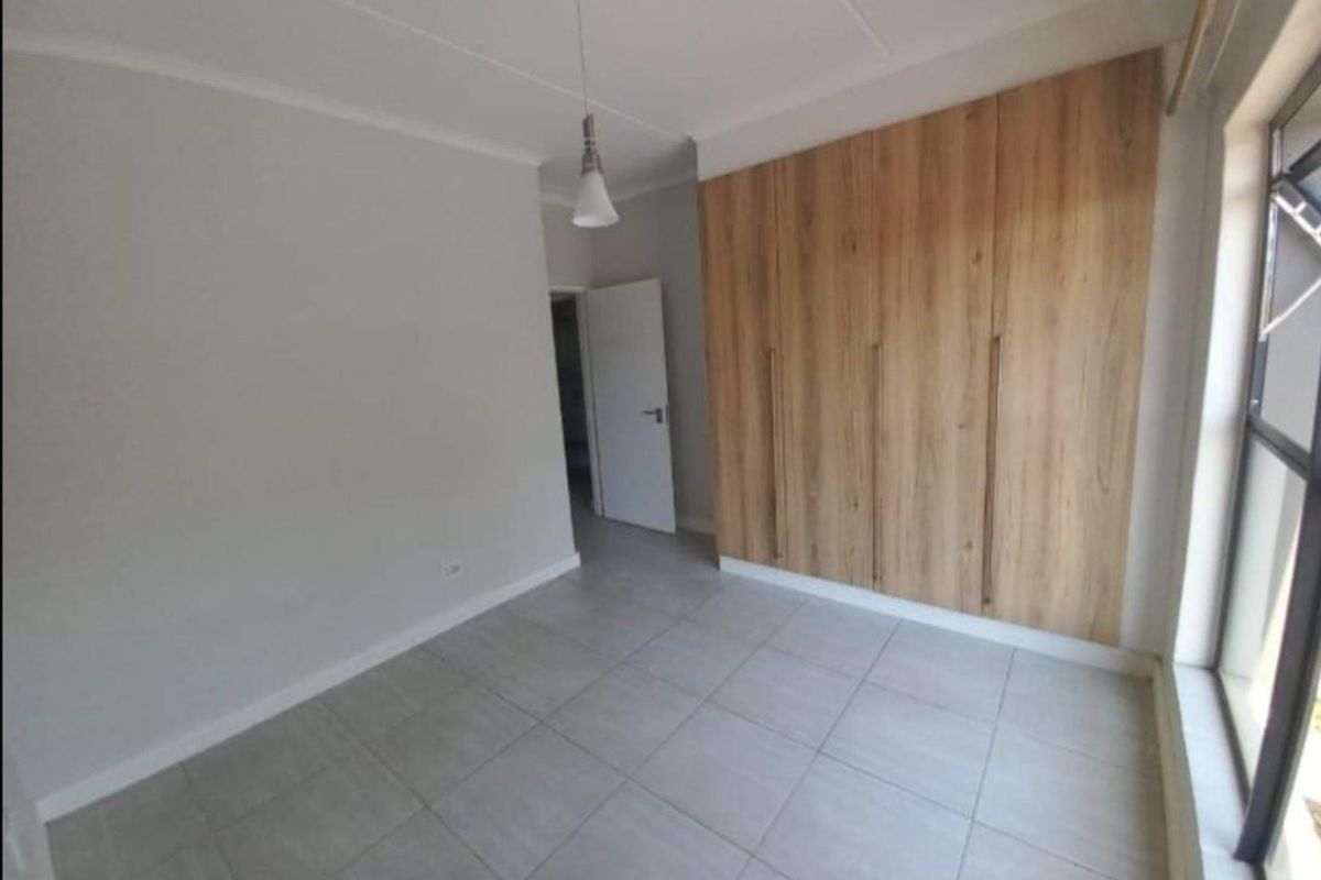 A bright, empty bedroom featuring modern wood-finish built-in cupboards, neutral tiled flooring, and a clean, minimalist aesthetic.