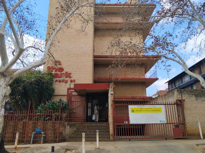 Front elevation of The Park apartment building featuring brick facade, multiple balconies, and secure gated entrance with perimeter fencing.