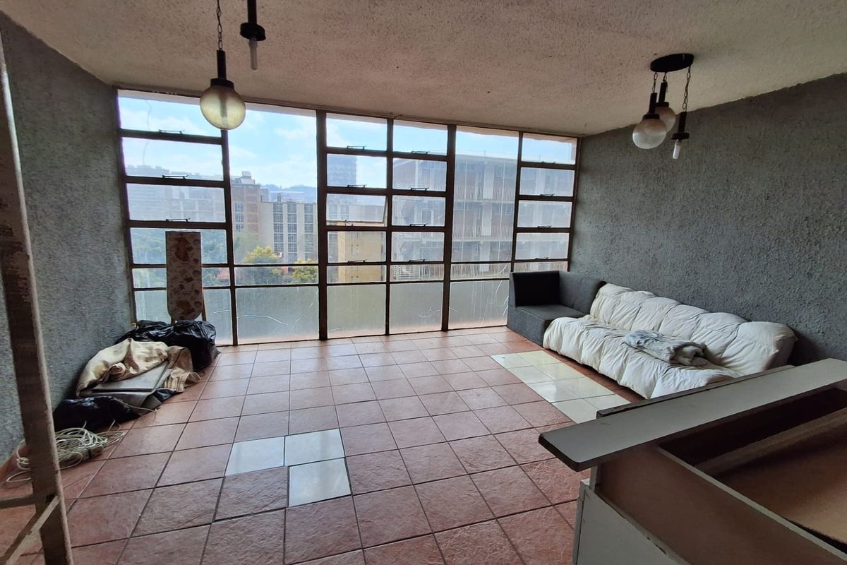 Spacious living area featuring large metal-framed windows with city views, tiled flooring, and pendant lighting, offering significant potential for renovation and modernization.