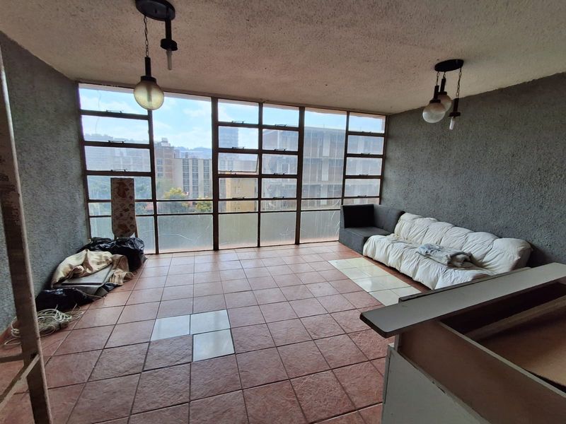 Spacious living area featuring large metal-framed windows with city views, tiled flooring, and pendant lighting, offering significant potential for renovation and modernization.