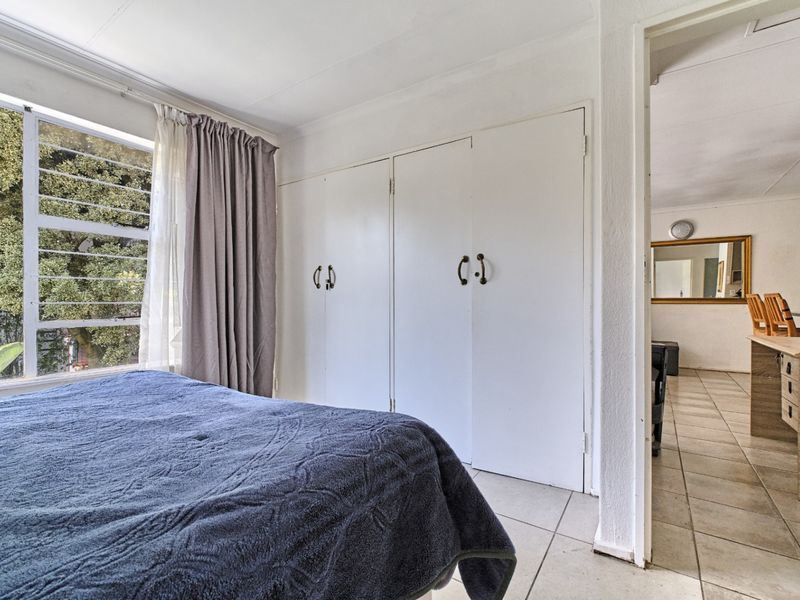 A bedroom featuring tiled flooring, built-in cupboards, and a large window, with an adjacent view into the living area.