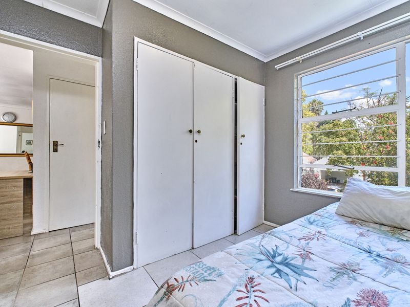A bedroom featuring built-in cupboards, tiled flooring, and large windows with security bars, offering a view of the surrounding greenery.
