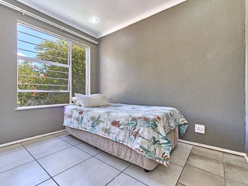 A compact bedroom featuring tiled flooring, a large window with security bars, and neutral grey walls.