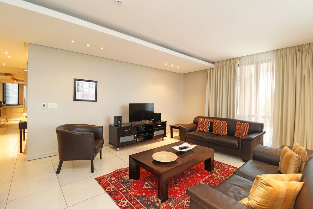 A spacious living room featuring a modern drop ceiling with recessed lighting, neutral tiled floors, and large windows with floor-to-ceiling curtains.