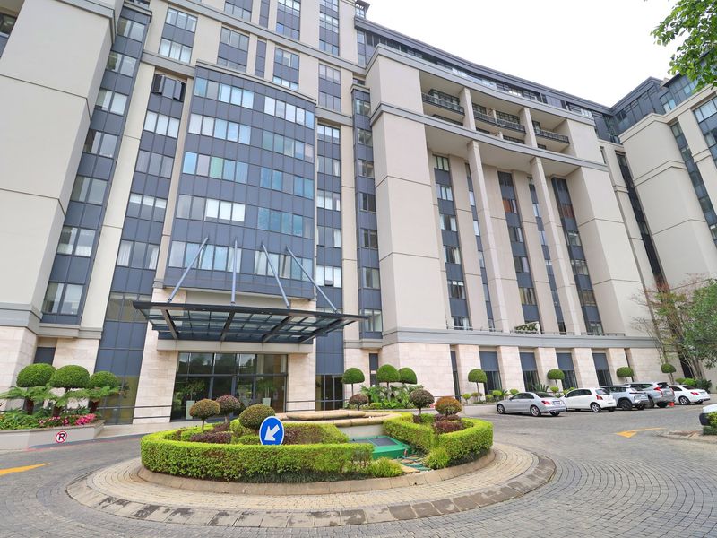 A modern apartment building exterior featuring a grand entrance with a glass canopy, stone facade, and a landscaped circular driveway.