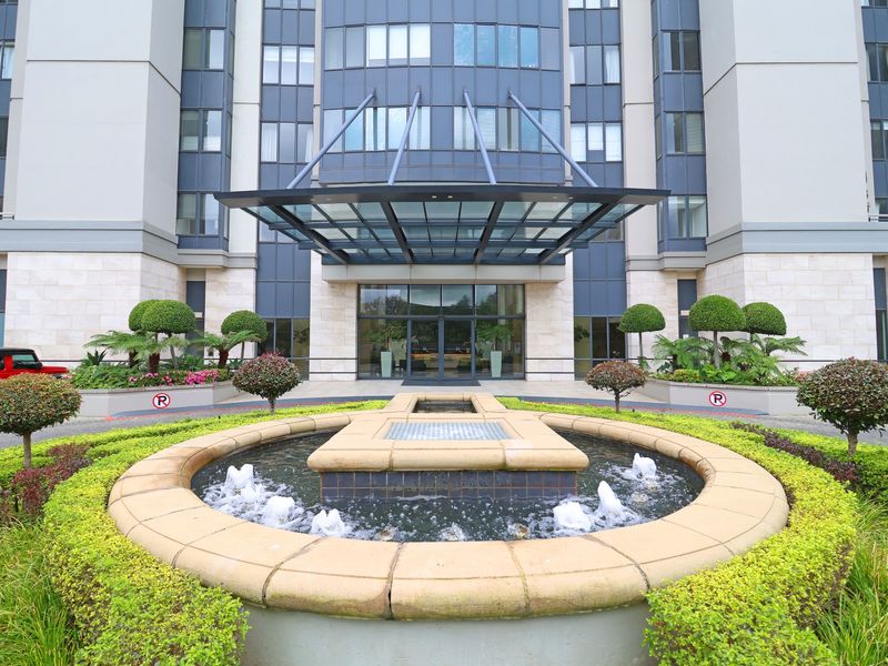 The front entrance of the apartment building features a modern glass canopy, a central water fountain, and well-maintained landscaped gardens.