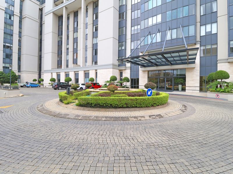 The front entrance of the apartment complex features a paved circular driveway, landscaped garden roundabout, and a modern glass canopy over the main entry.