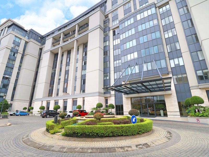 Modern apartment building exterior featuring a glass entrance canopy, stone facade, and a circular paved driveway with landscaped garden.