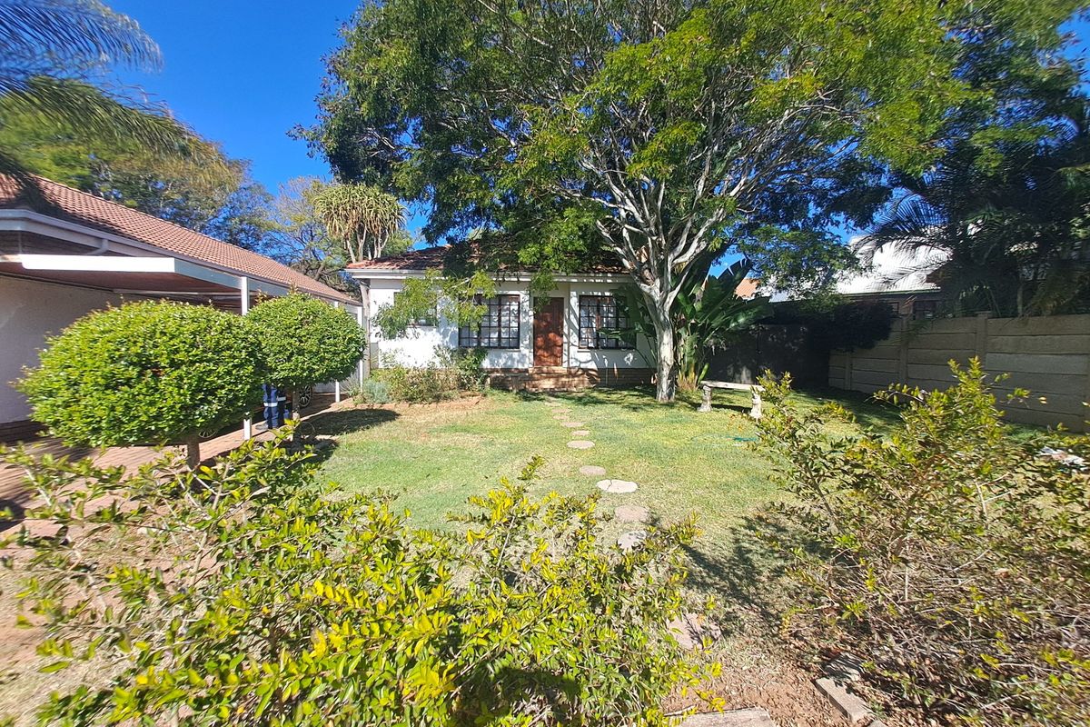 A rear view of the house featuring a well-maintained garden, a covered patio area, and mature trees providing shade.
