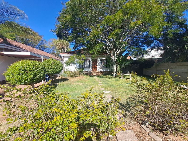 A rear view of the house featuring a well-maintained garden, a covered patio area, and mature trees providing shade.