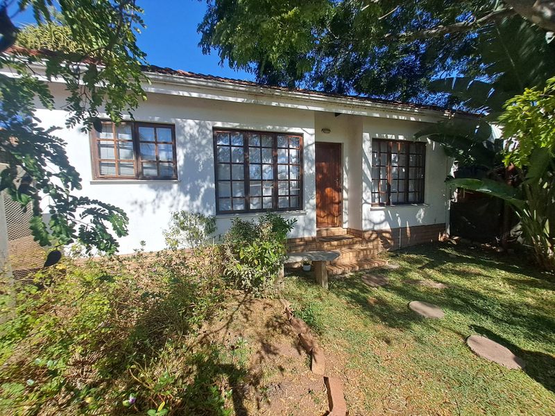 Front elevation of a single-story house featuring timber-framed windows, a tiled roof, and a brick entrance area surrounded by a garden.