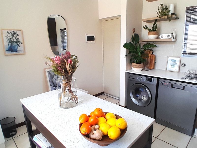 A well-maintained kitchen area featuring a central island with stone countertop, tiled flooring, floating shelving, and integrated laundry and dishwasher appliances.