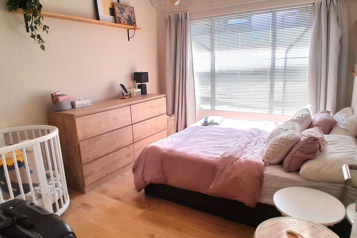 A well-lit bedroom featuring laminate flooring, a large window with venetian blinds, and a floating wall shelf.