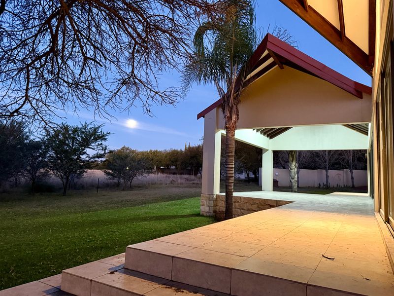 A spacious covered patio featuring tiled flooring, exposed timber beams, and stone cladding, overlooking a well-maintained garden at twilight.