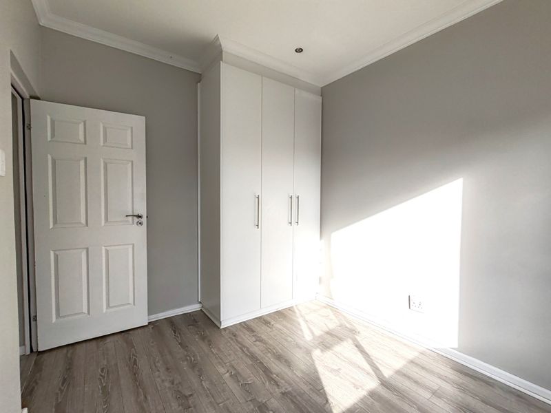 A bright, empty bedroom featuring modern laminate flooring, neutral grey walls, and white built-in cupboards with clean lines.