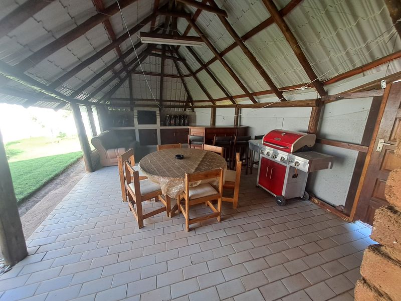 Covered outdoor patio area featuring a thatched roof, exposed timber rafters, tiled flooring, and a built-in braai, offering a rustic entertainment space.