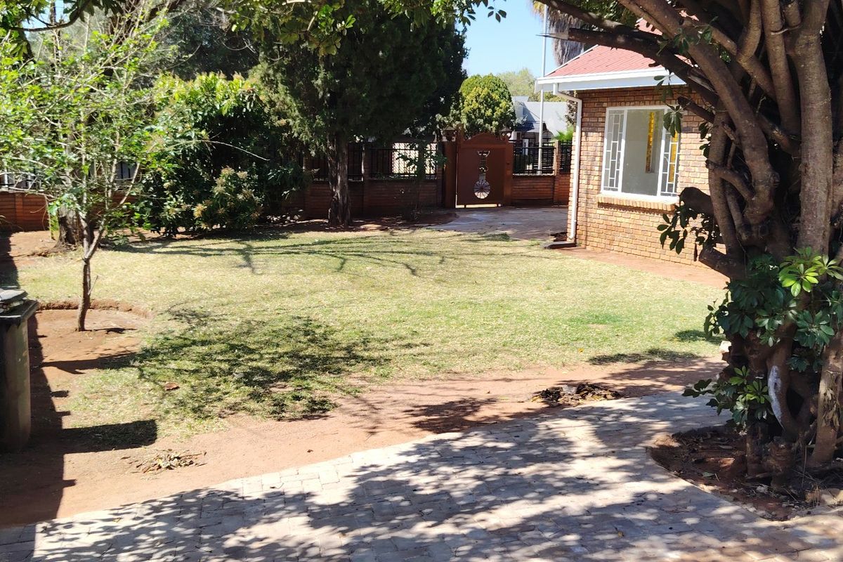A view of the rear garden featuring a brick exterior wall, paved walkway, and established trees with a lawn area.