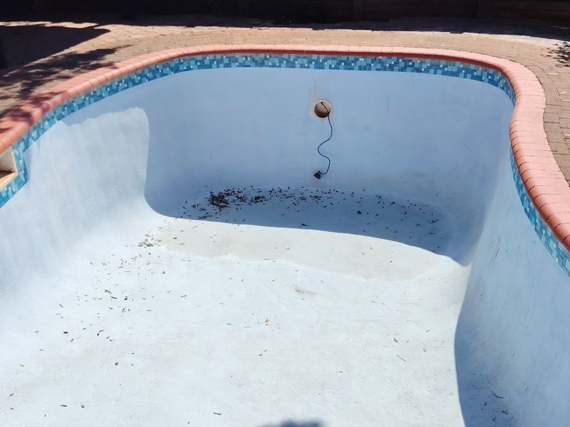 An empty swimming pool featuring a mosaic tile border and brick paved surround, currently requiring maintenance and cleaning.