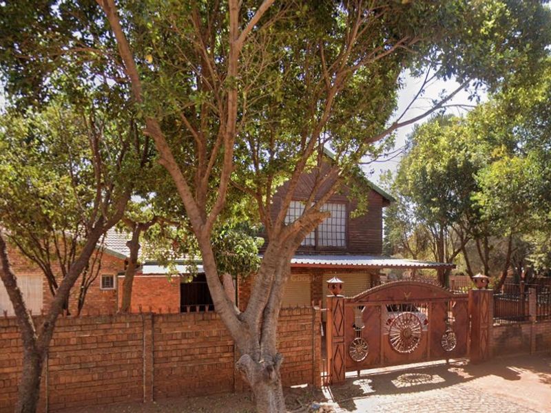 Front view of a face brick house featuring a decorative metal gate, brick boundary wall, and mature trees providing shade to the property entrance.
