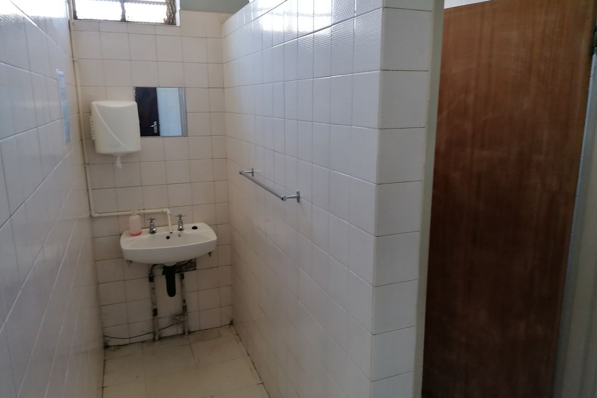 A functional commercial bathroom featuring white tiled walls, a wall-mounted basin, and a timber door, requiring cosmetic updates to improve its overall condition.