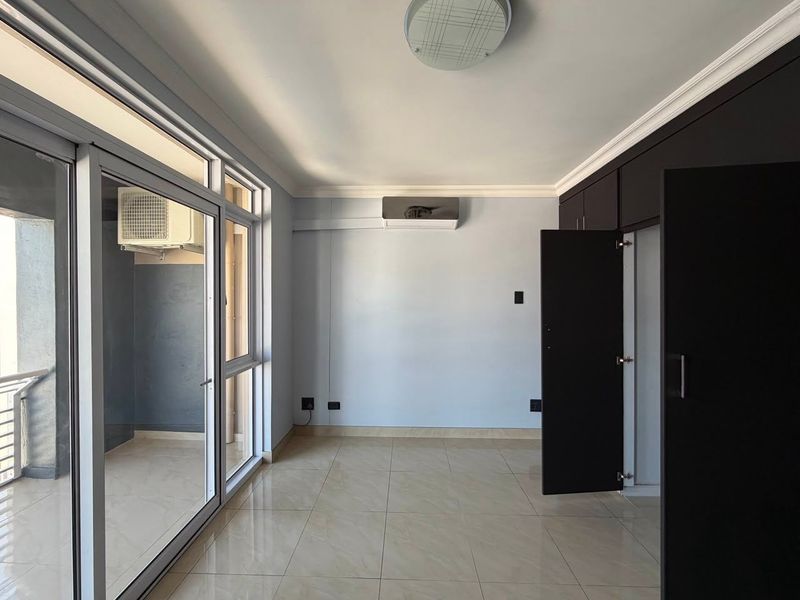 A bright, empty bedroom featuring tiled floors, built-in dark cabinetry, an air conditioning unit, and large sliding glass doors leading to a balcony.