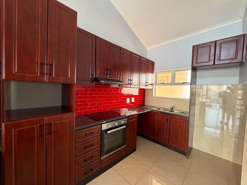 A clean, functional kitchen featuring dark wood cabinetry, granite countertops, a red tiled backsplash, and integrated appliances including an oven and hob.