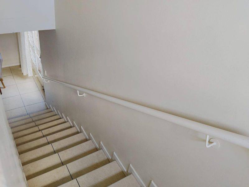 A clean, neutral-toned staircase featuring tiled steps and a simple white wall-mounted handrail, leading down to a tiled landing area.