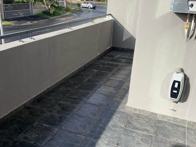 A clean, tiled balcony featuring a metal balustrade, external gas geyser, and power point, offering an open view of the surrounding street area.