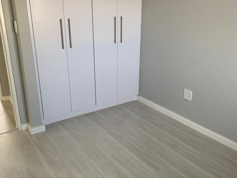 A clean, empty bedroom featuring light-toned laminate flooring, neutral grey walls, and modern built-in cupboards with sleek handles.