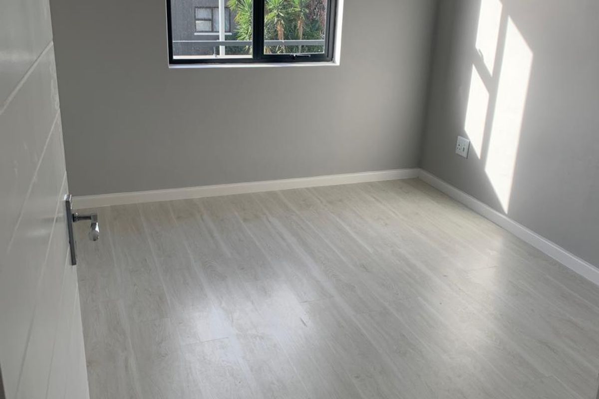 A bright, empty bedroom featuring light-toned laminate flooring, neutral grey walls, and a modern black aluminium window frame.