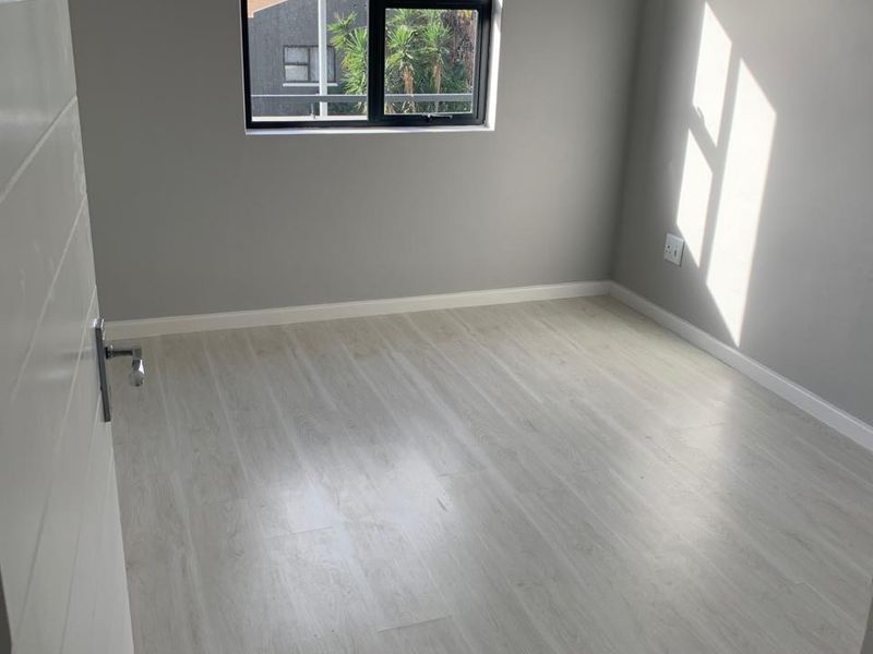 A bright, empty bedroom featuring light-toned laminate flooring, neutral grey walls, and a modern black aluminium window frame.