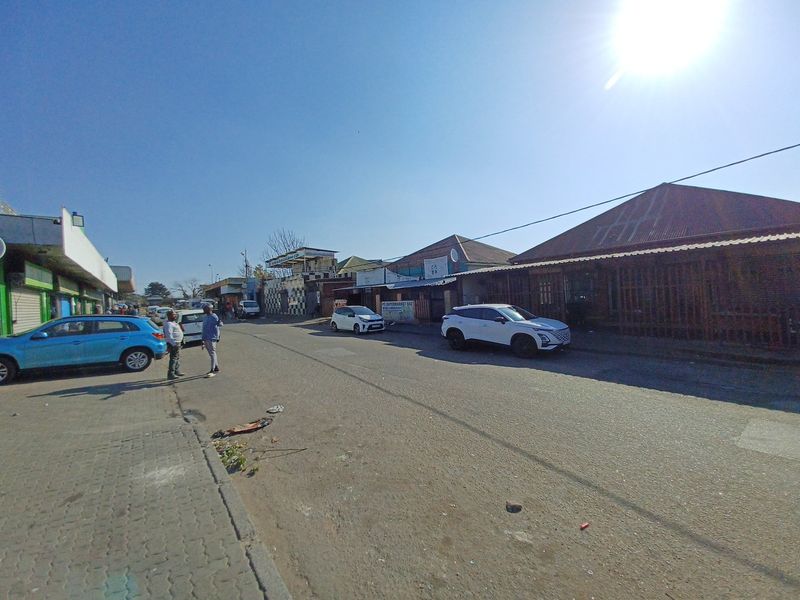 A street-level view of a commercial area featuring asphalt roads, paved sidewalks, and various storefronts with corrugated metal roofing.