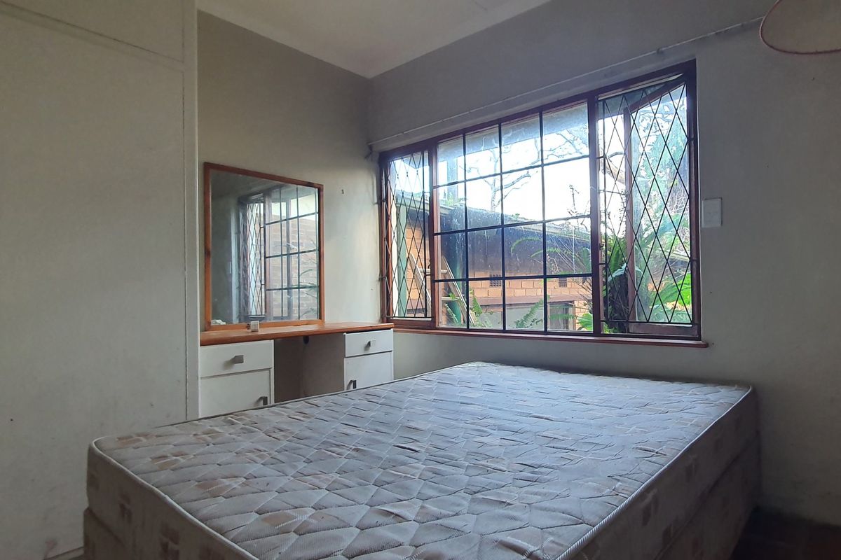 A bedroom featuring built-in wardrobes, a vanity unit, and large windows with security bars, offering a functional space in need of cosmetic updates.