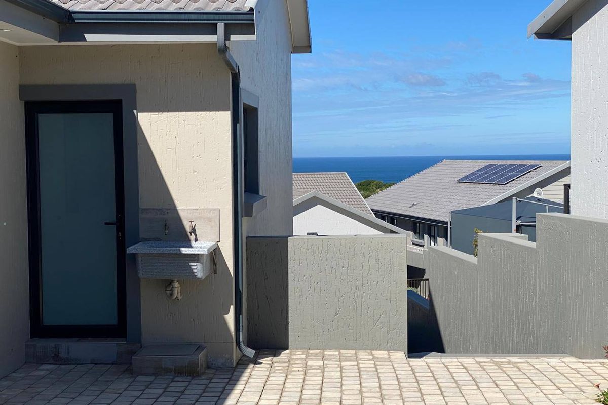 Rear exterior view featuring a paved patio, external utility sink, and distant ocean views with neighbouring properties visible.