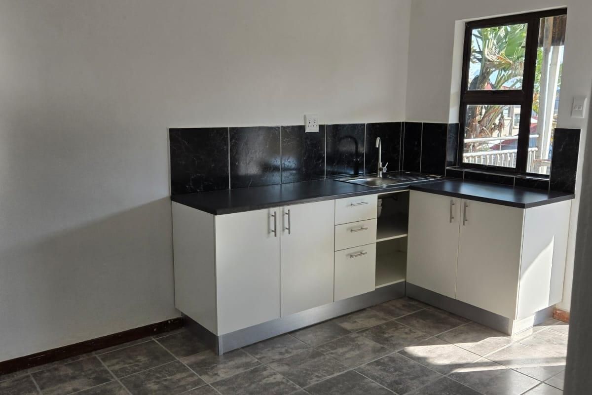 A clean, empty kitchen featuring white built-in cabinetry, dark countertops, a tiled backsplash, and grey tiled flooring with natural light from the window.