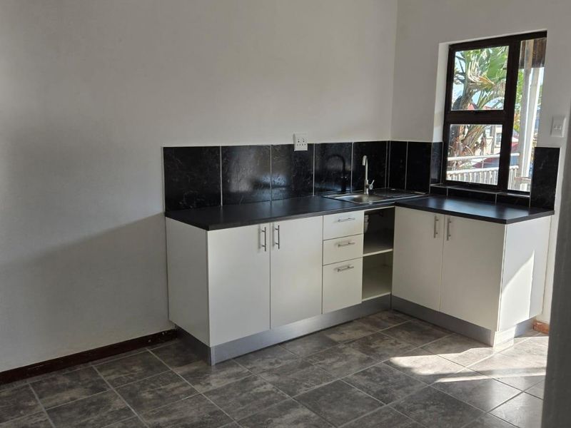 A clean, empty kitchen featuring white built-in cabinetry, dark countertops, a tiled backsplash, and grey tiled flooring with natural light from the window.