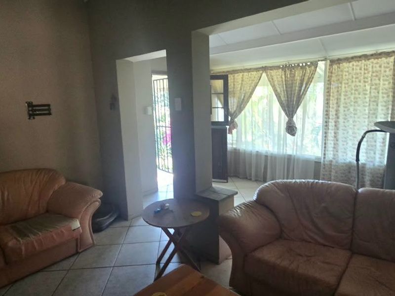 A living room area featuring tiled flooring and large windows, providing access to an adjacent sunroom or patio space through a doorway.