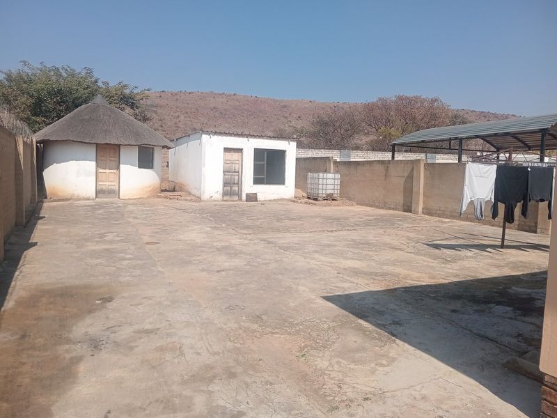 A spacious rear yard featuring a traditional thatched rondavel, a separate outbuilding, and a concrete paved area enclosed by perimeter walls.