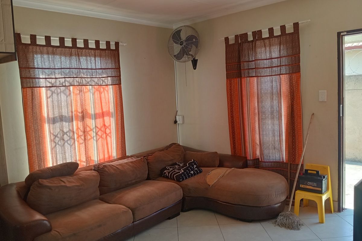 A living room area featuring tiled flooring, wall-mounted fan, and dual windows with curtain rods, providing a functional space for relaxation.