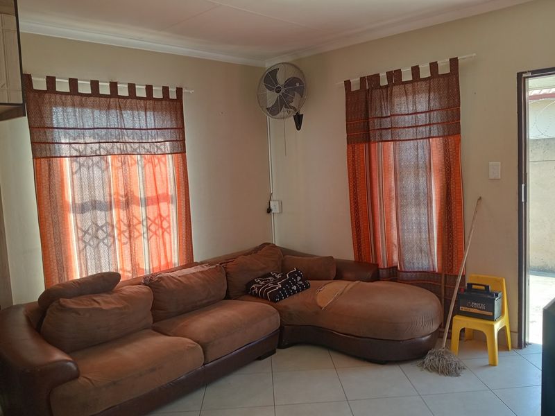 A living room area featuring tiled flooring, wall-mounted fan, and dual windows with curtain rods, providing a functional space for relaxation.
