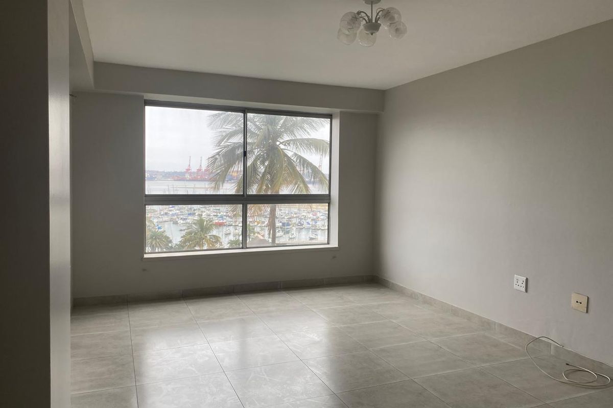 A bright, empty living room featuring large windows with a scenic harbor view and neutral tiled flooring.